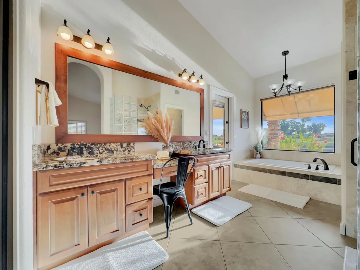 A serene bathroom with a freestanding bathtub beside a large window, a granite countertop with dual sinks, a spacious mirror, and natural light pouring across the room. Greenery adds a refreshing touch.