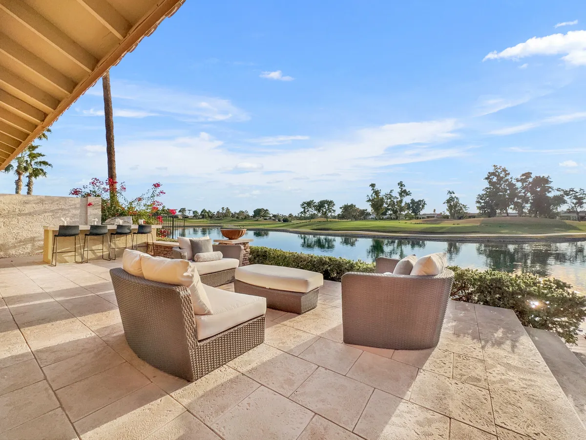 A beautifully arranged outdoor seating area on the waterfront, surrounded by lush greenery. The reflective water surface creates a tranquil ambiance, while the distant golf course hints at resort-style amenities. The scene is perfect for relaxing with friends or enjoying a quiet morning coffee by the lake.