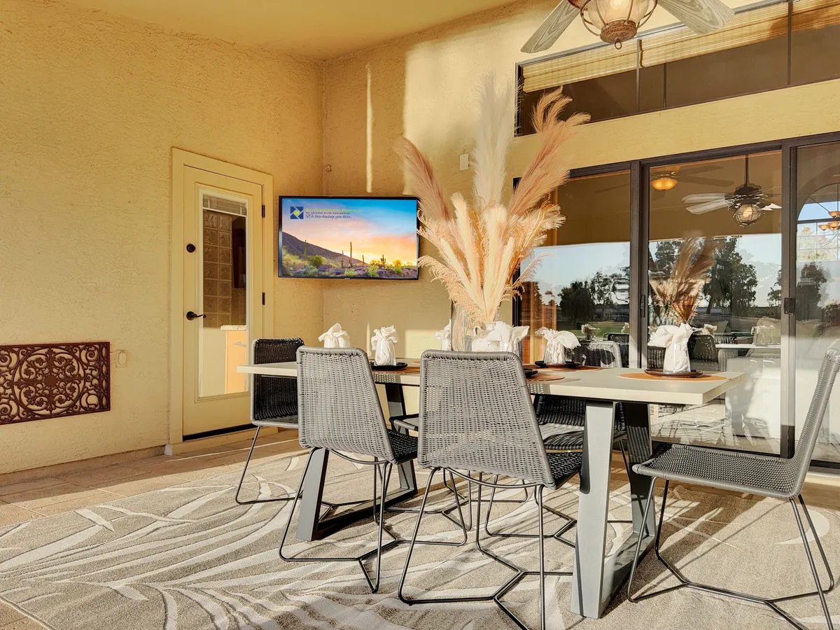 A stylish outdoor dining area featuring a table with a decorative centerpiece, pampas grass arrangement, and a television displaying a scenic view. Comfortable, modern chairs surround the table, and the setup is illuminated by natural light, suggesting an open and welcoming space for gatherings. The ambiance is relaxed yet festive, ideal for both daytime meals and evening celebrations.