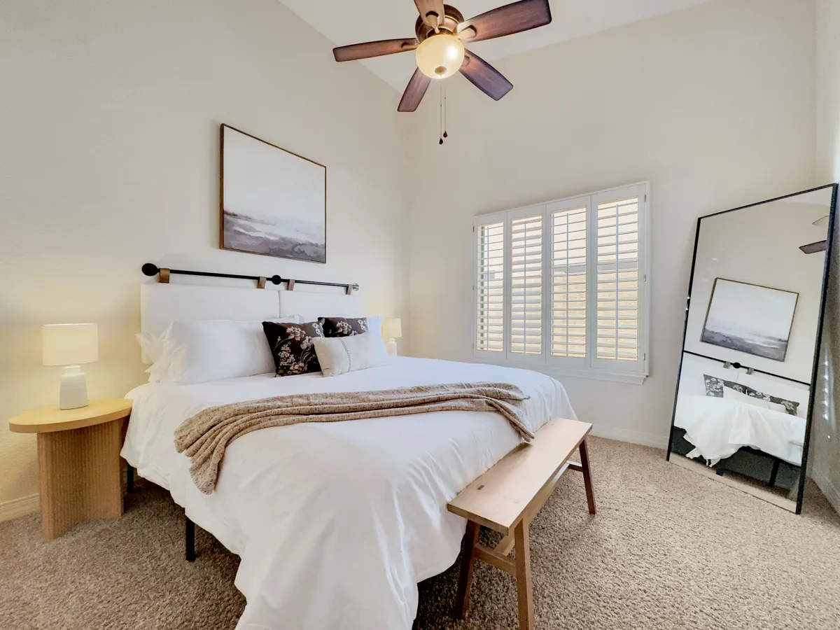 A bright, serene bedroom with a bed dressed in crisp white linens, a large mirror, soft lamp lighting, and gentle artwork. The space feels peaceful and inviting, ideal for restful sleep or quiet mornings.
