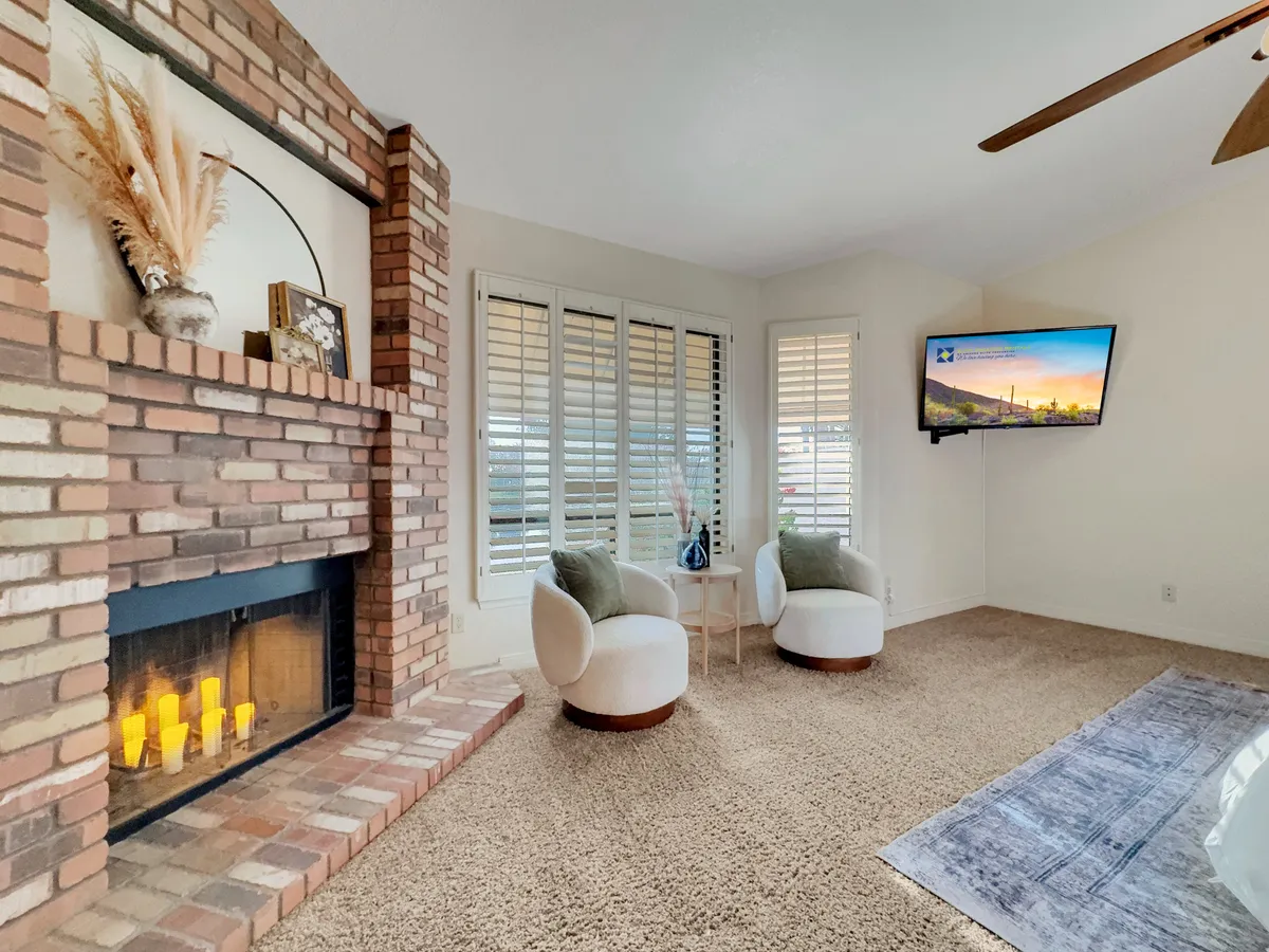 A cozy main living area featuring a brick fireplace, plush and comfortable seating, and a TV mounted on the wall. Large picture windows let in abundant natural light and offer glimpses of tranquil exterior views. Decorative plants and thoughtfully chosen furnishings elevate the atmosphere, making the space perfect for relaxing, socializing, or movie nights.