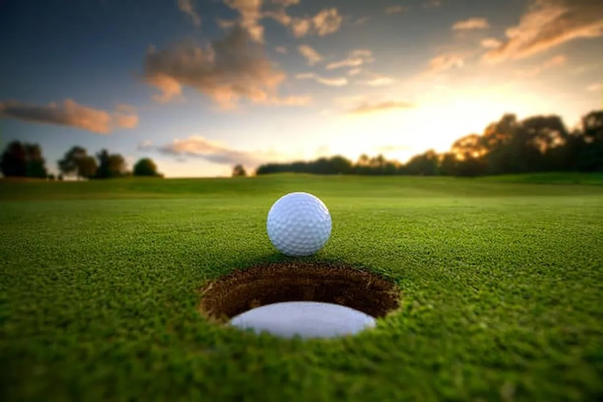 A close-up view of a golf ball poised at the edge of a hole on a pristine, lush green fairway. The scene is bathed in natural light, with the texture of the grass and the open, peaceful ambiance of the golf course clearly visible.
