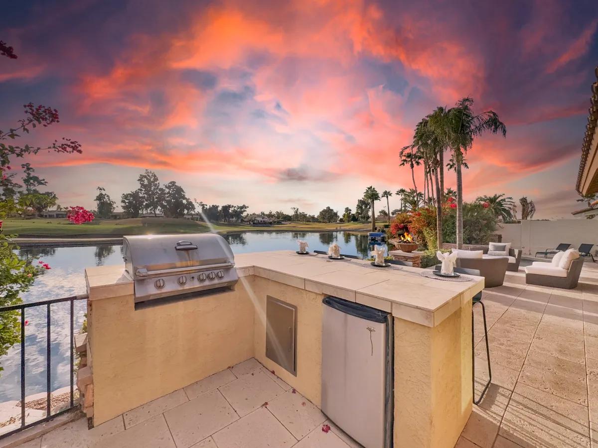 An inviting outdoor kitchen area featuring a built-in gas BBQ, comfortable seating, and a panoramic lake view under a colorful sunset sky. The space is ideal for grilling and outdoor dining.