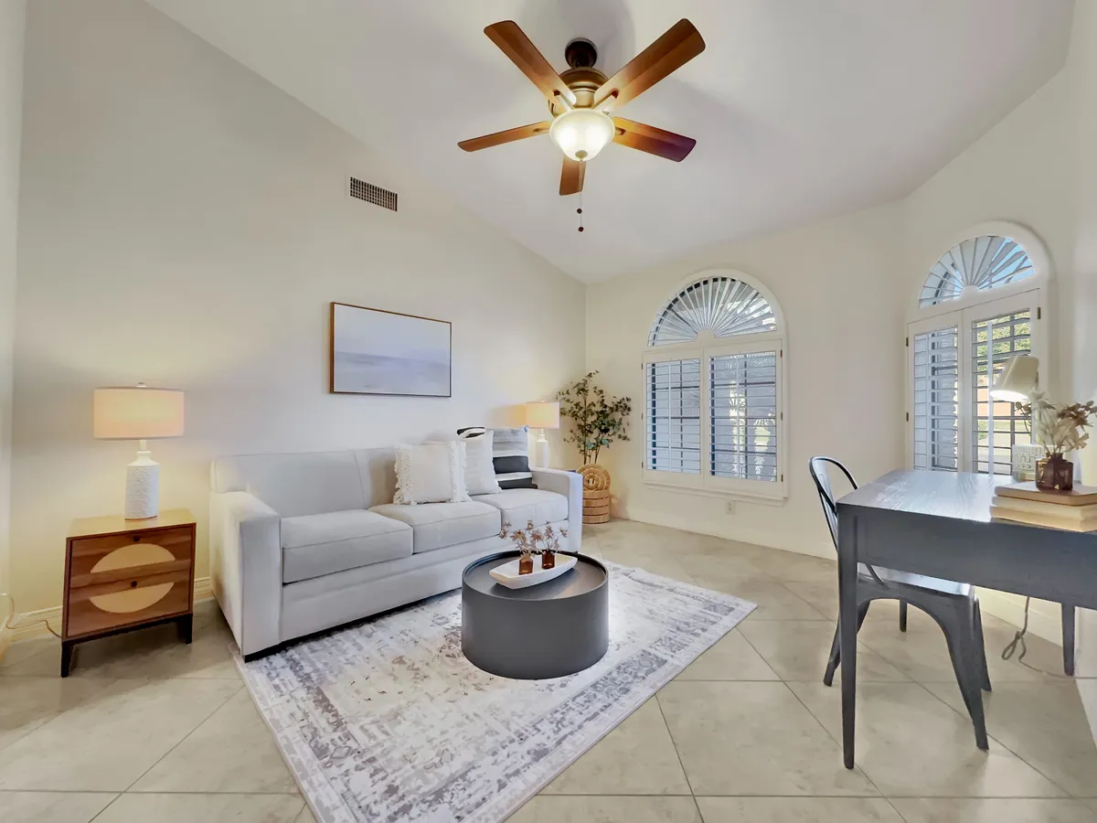A stylish, multi-functional office or flex space with a comfortable sofa, decorative rug, an elegant desk and chair, and a ceiling fan. Natural light streams in, making it inviting both for productivity or as a cozy nook for relaxation or extra sleeping space.