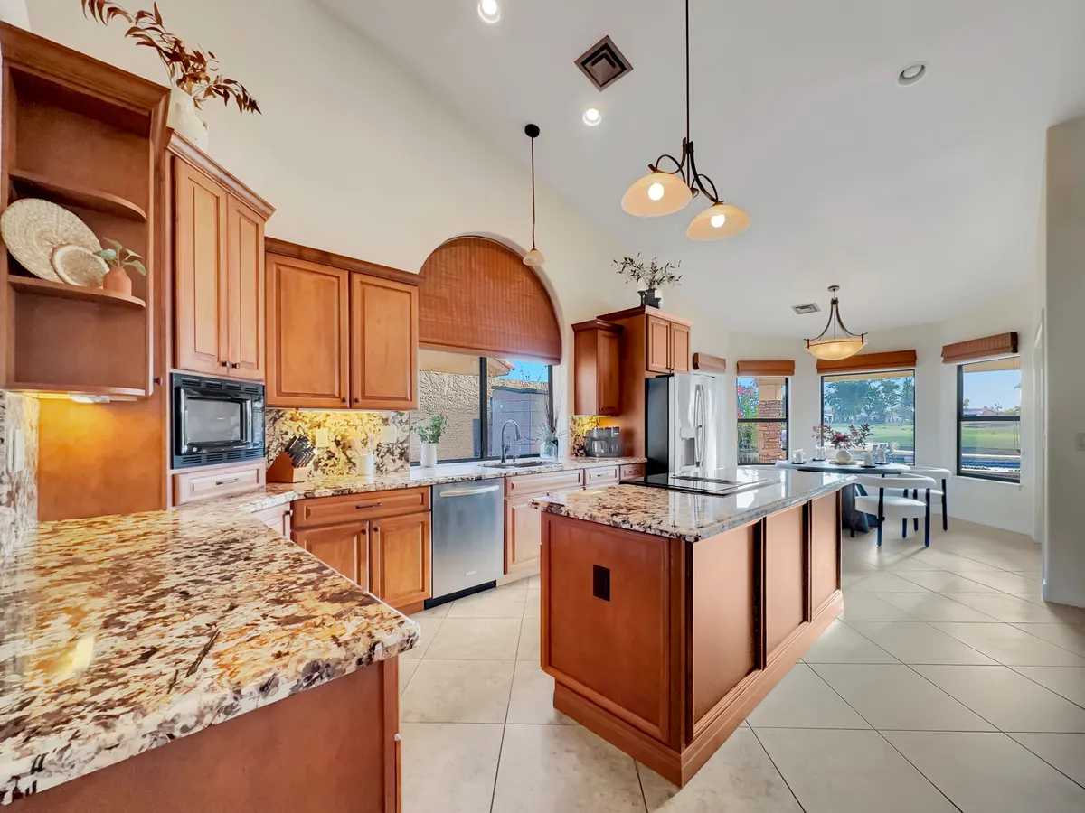 A warmly-lit kitchen featuring granite countertops, wood cabinetry, a spacious island with seating, and large windows that invite natural light. The space is clearly designed for both cooking and socializing, with ample room for multiple people to gather around the island or work together.