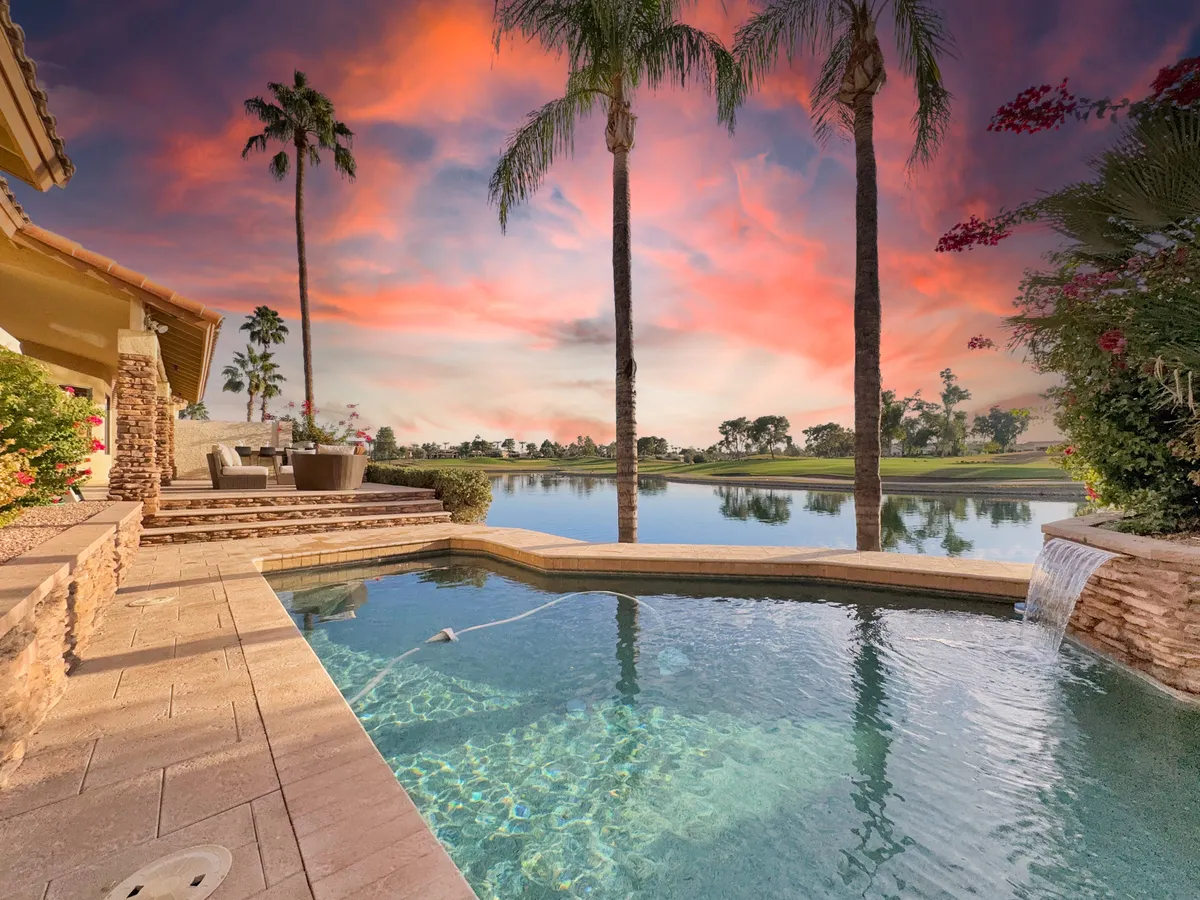 A serene backyard pool framed by palm trees, with a stunning sunset sky reflected on the water. The poolside area is designed for both relaxation and outdoor entertaining, with seating and an unobstructed view of the lake beyond.