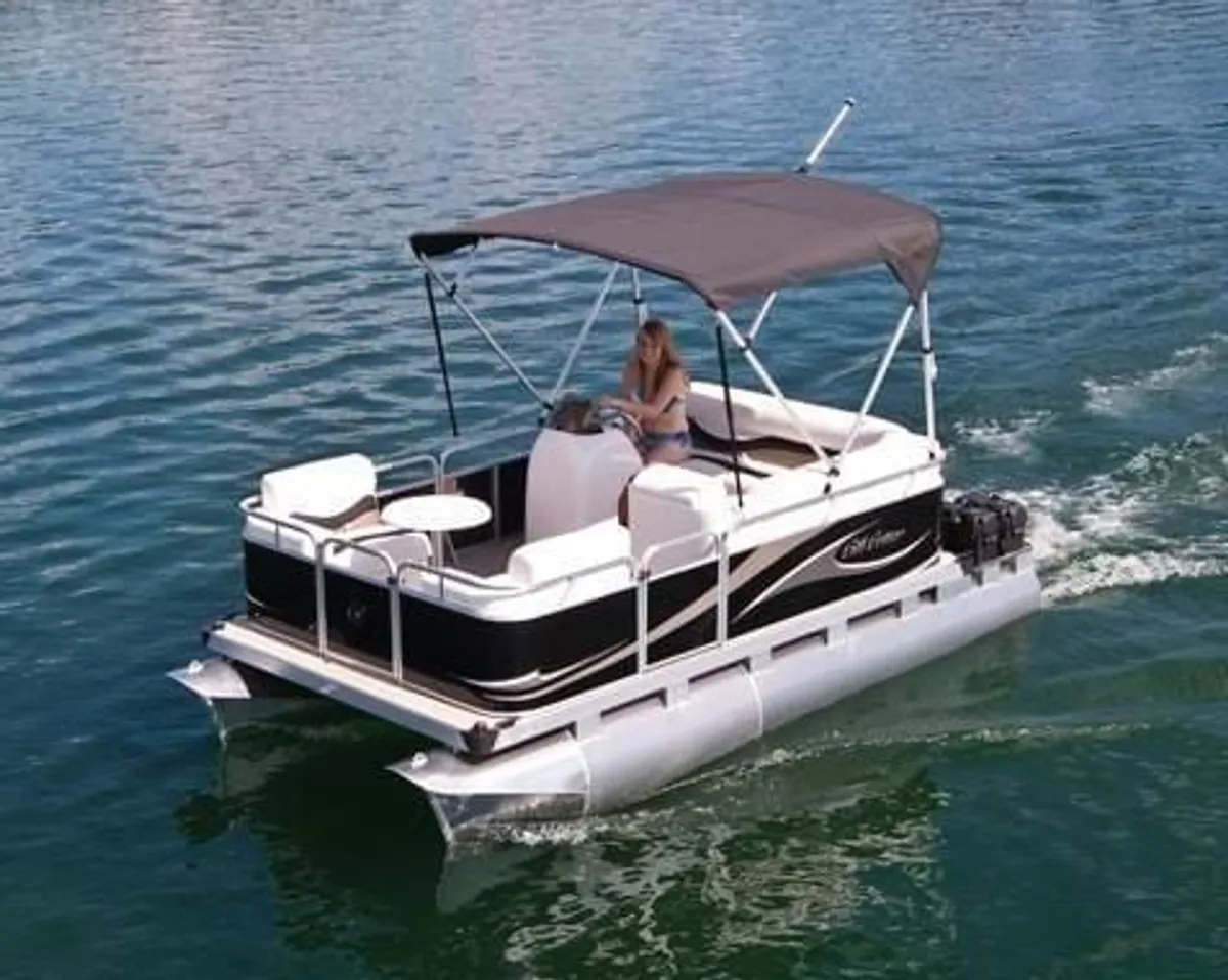 A pontoon boat gently cruising on the water, captained by a woman under a canopy, with calm lake views and a sense of leisurely adventure.